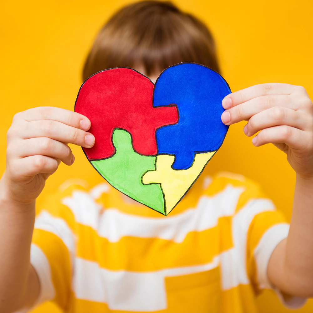 Dłonie chłopca trzymające kolorowe puzzle w kształcie serca na tle żółtym. Światowy Dzień Świadomości Autyzmu, koncepcja zaburzeń ze spektrum autyzmu.