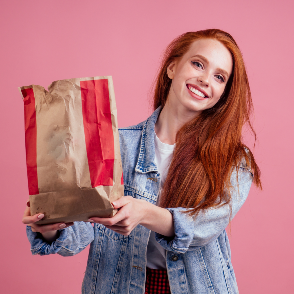Uśmiechnięta kobieta trzyma papierową torbę przypominającą barwy KFC z zakupami na różowym tle